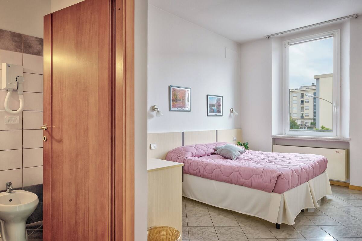 Quarto com cama, mesa de cabeceira, janela e vista para edifícios.