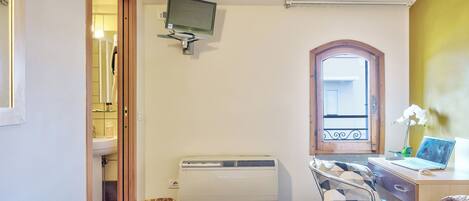 Un dormitorio con cama, escritorio con computadora portátil, ventana y baño visible a través de una puerta abierta.