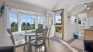 Habitación luminosa con una mesa, sillas, ventanas amplias, una puerta que da al exterior y una zona de cocina.