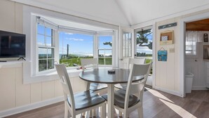 Un coin repas avec une table ronde et des chaises blanches, de grandes fenêtres donnant sur la mer et un téléviseur posé sur un meuble.