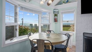 Un comedor con una mesa redonda y sillas, ventanas grandes con persianas, una lámpara de araña y vista al mar.