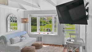 Ein gemütliches Zimmer mit weißem Sofa, einem großen Fenster mit Blick auf Grünflächen, einem an der Wand befestigten Flachbildfernseher und einem kleinen Tisch mit Stuhl.