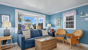 Una sala de estar con un sofá azul, sillas de mimbre, una mesa de centro de vidrio y vistas al jardín a través de ventanas amplias.