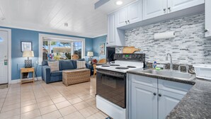 Cocina moderna con armarios blancos, estufa y lavabo. En la sala de estar, hay un sofá azul y una cesta de mimbre.