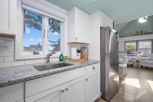 A kitchen with white cabinets, a granite countertop, a stainless steel refrigerator, and a window with a view of trees.