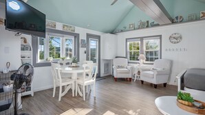 A spacious living room with a high ceiling, large windows, and a view of the outdoors. There are white furniture pieces, including a dining table and chairs, and a seating area with two armchairs. A flat-screen TV is mounted on the wall, and there are decorative items on the shelves.