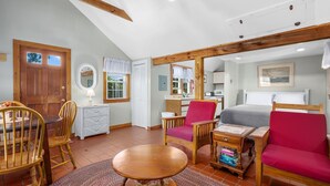A cozy room with a wooden ceiling, a bed with a red headboard, a dining table with chairs, a small kitchen area, and a window with curtains.