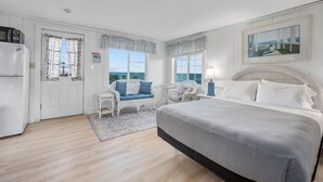 A bedroom with a bed, a chair, a small table, a refrigerator, and a window with a view of the sea.