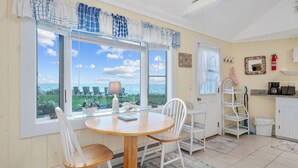 Un coin repas avec une table ronde en bois et des chaises blanches, de grandes fenêtres avec des rideaux, une vue sur la mer, et un espace cuisine avec des meubles blancs et une machine à café.
