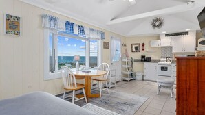 Une cuisine chaleureuse avec des meubles blancs, une cuisinière et un four à micro-ondes. On y trouve un coin repas avec une table et des chaises, et une vue sur la mer grâce à de grandes fenêtres.
