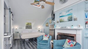 A bedroom with a bed, a dresser, a nightstand, a window, a ceiling fan, and a fireplace. - Lost at Seaside Too – Salt, Sand, Serenity (South Yarmouth)