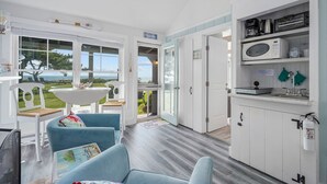 A bright, open-plan living area with a view of the sea, a kitchen with white cabinets, and a dining area with blue chairs. - Lost at Seaside Too – Salt, Sand, Serenity (South Yarmouth)