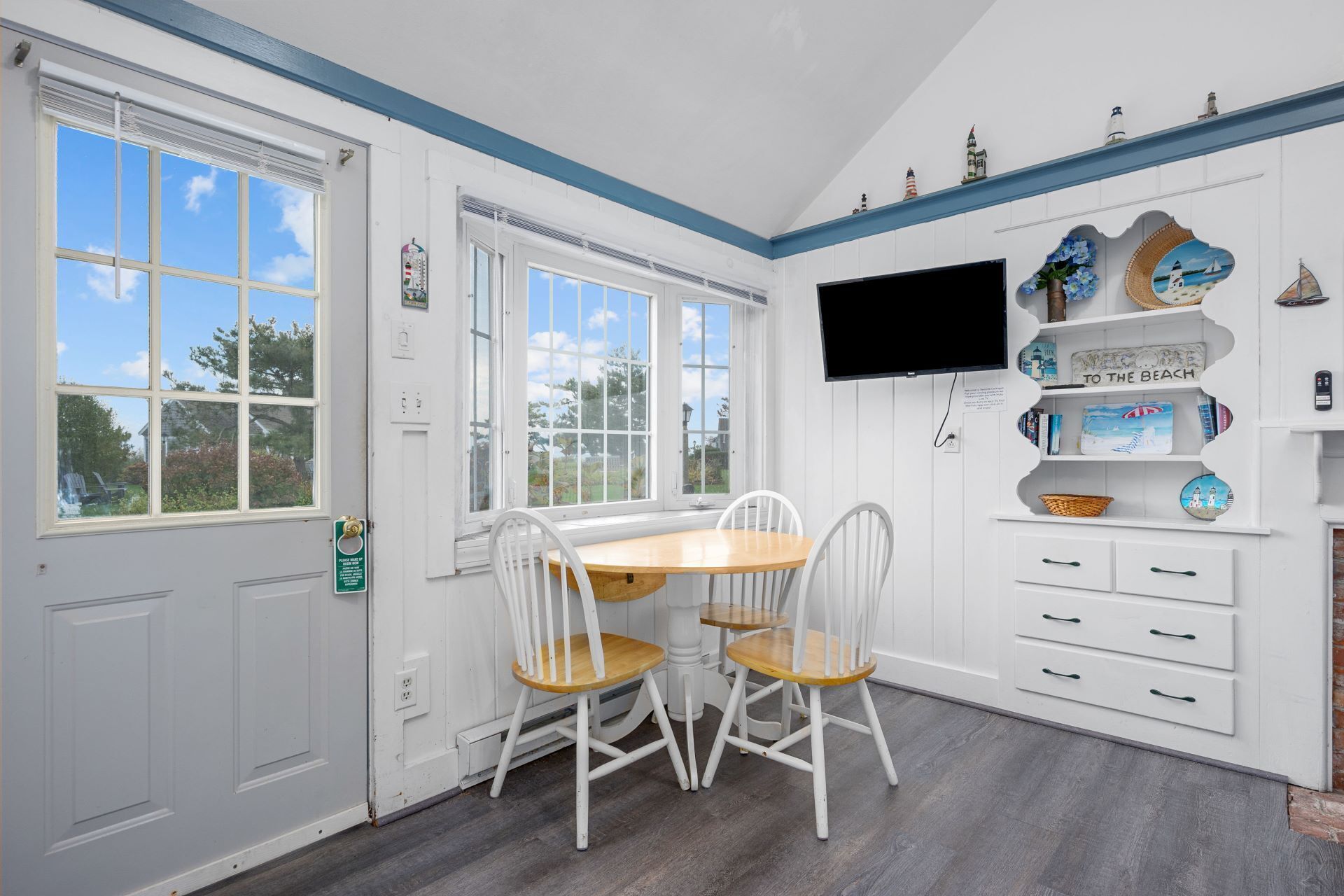 A bright room with a wooden table and white chairs, a flat-screen TV, and decorative items on shelves.
