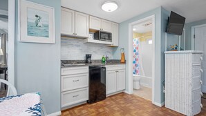 Une cuisine compacte avec des meubles blancs, un four à micro-ondes et un lave-vaisselle. On peut voir un plancher en bois et une salle de bain à l’arrière-plan.