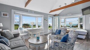 Een ruime woonkamer met uitzicht op zee, een eethoek en een gezellig zithoek met een gestreept sofa en een klein rond tafeltje.