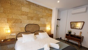 A neatly made bed with white linens and a rolled towel, a wooden nightstand with a lamp, and a stone wall behind the bed.