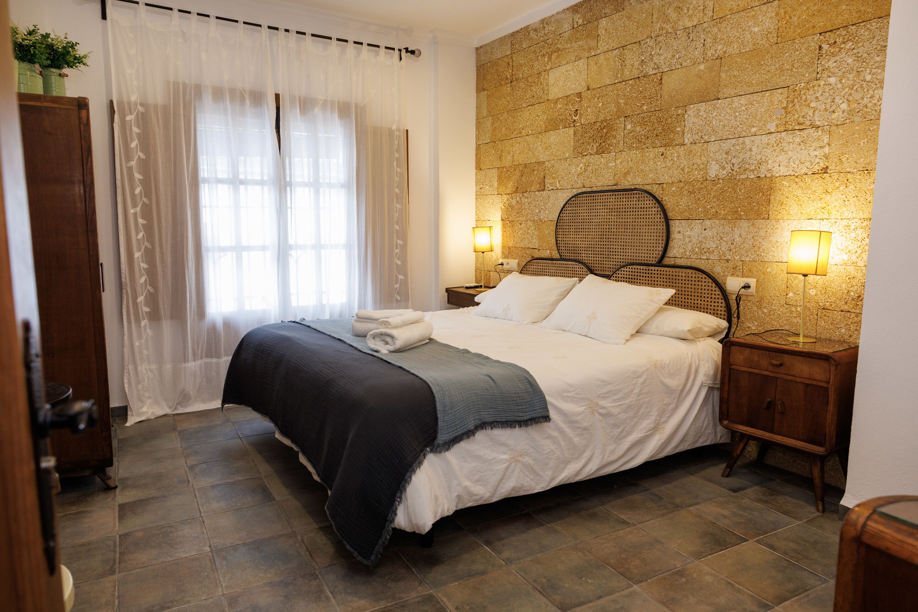 A bedroom with a bed, two bedside tables with lamps, a wooden headboard, and a stone wall.
