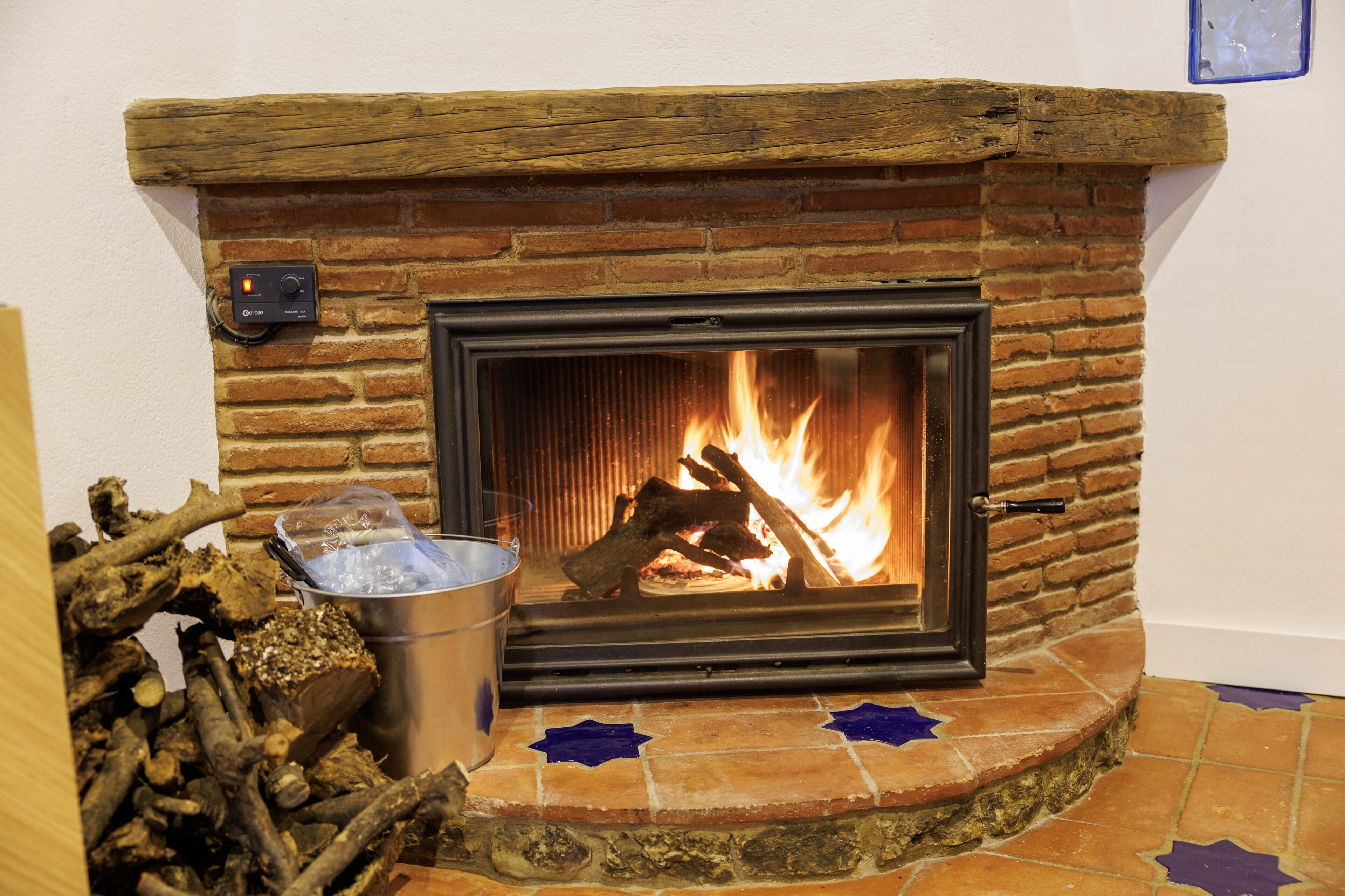 A fireplace with a brick surround and a metal grate, with logs and a bucket of kindling nearby.