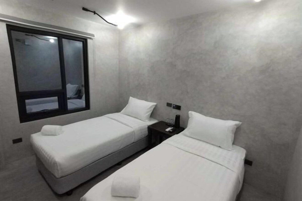 A small, modern hotel room with two single beds, a nightstand, a window, and a wall-mounted light fixture.