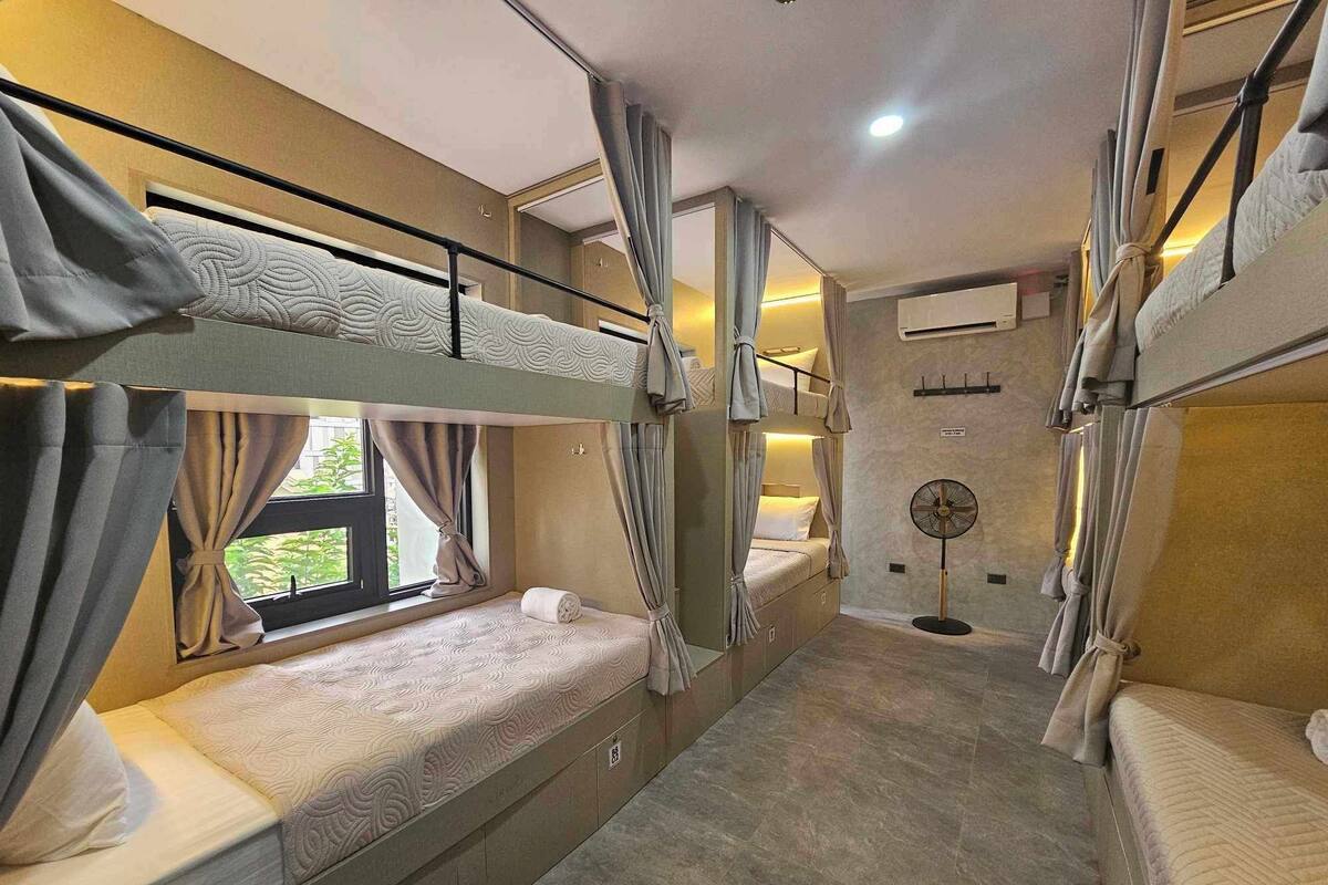 A bunk bed room with curtains, a standing fan, and a window with a view.