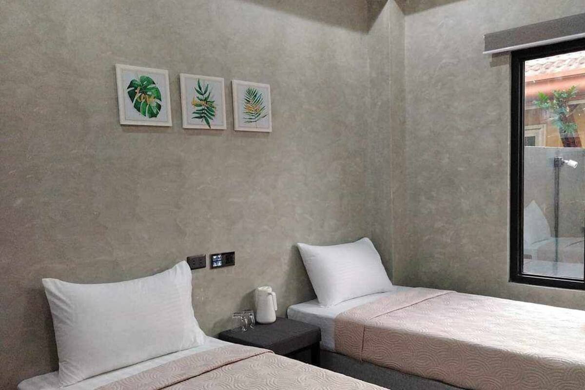 A hotel room with two beds, a small table, a window, and three framed botanical prints on the wall.