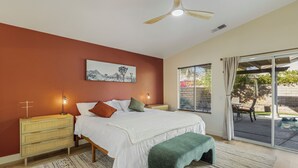 A bedroom with a bed, bedside tables, a nightstand, a bench, a ceiling fan, a window with blinds, and a view of the outdoors.
