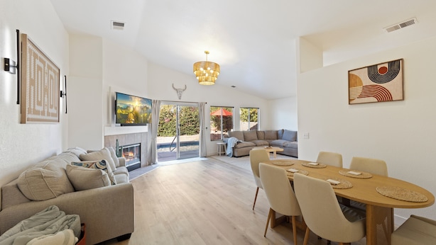 A spacious living room with a fireplace, a dining area with a wooden table and chairs, and a large window with a view of the outdoors.