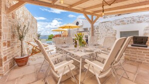 Un patio al aire libre con una mesa dispuesta para comer, rodeado de sillas de mimbre, una planta en maceta y vistas al mar.