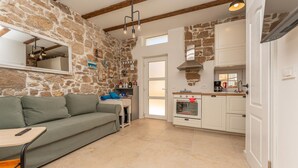 Una sala de estar acogedora con una pared de piedra, un sofá y una pequeña mesa. Hay una cocina con armarios blancos y una estufa.