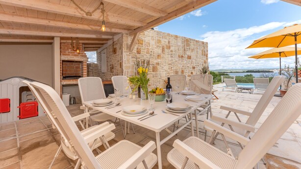 Un área de comedor al aire libre con un fondo de pared de piedra, muebles de mimbre blanco y vistas al mar.