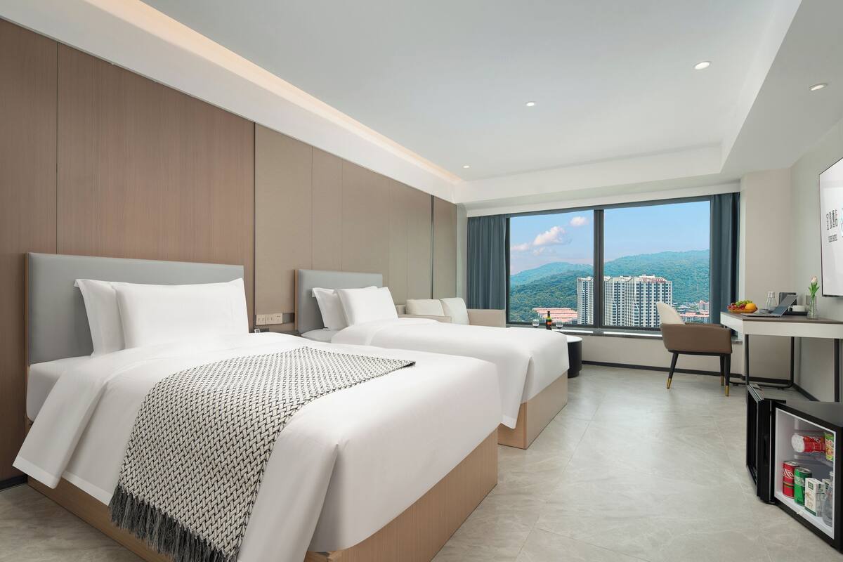 Habitación de hotel con dos camas, un escritorio y un ventanal grande con vistas a la ciudad.