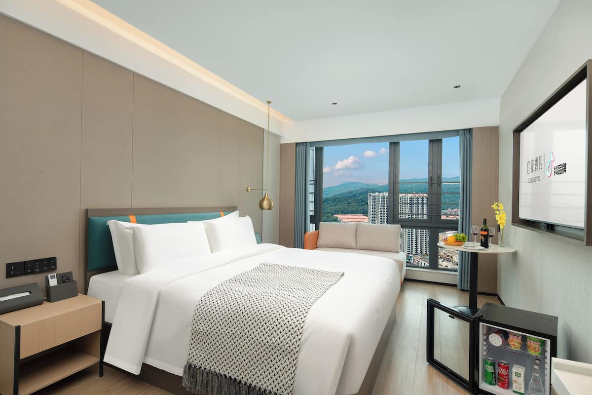 Habitación de hotel moderna con una cama grande, una mesita con una botella y flores, un mini refrigerador y vistas a la ciudad.