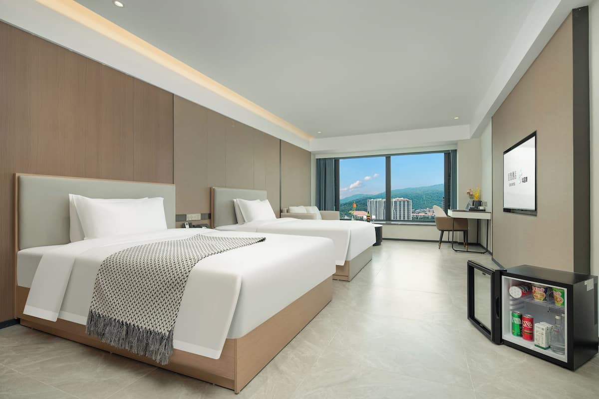 Habitación de hotel moderna con dos camas, un escritorio pequeño, un refrigerador y un ventanal grande con vistas a la ciudad.