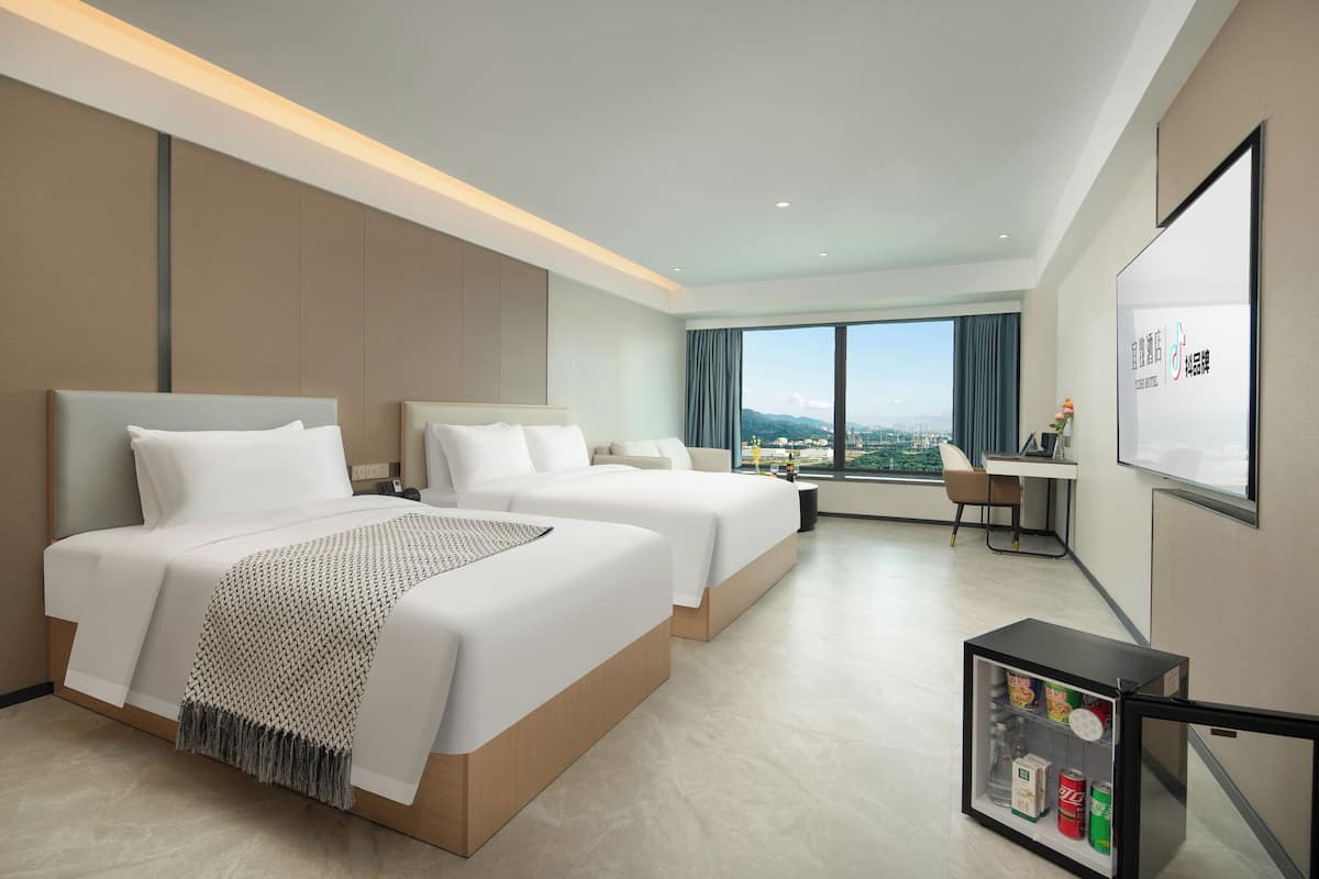 Habitación de hotel con dos camas, un escritorio, una silla, un refrigerador pequeño y un amplio ventanal con vistas a la ciudad.