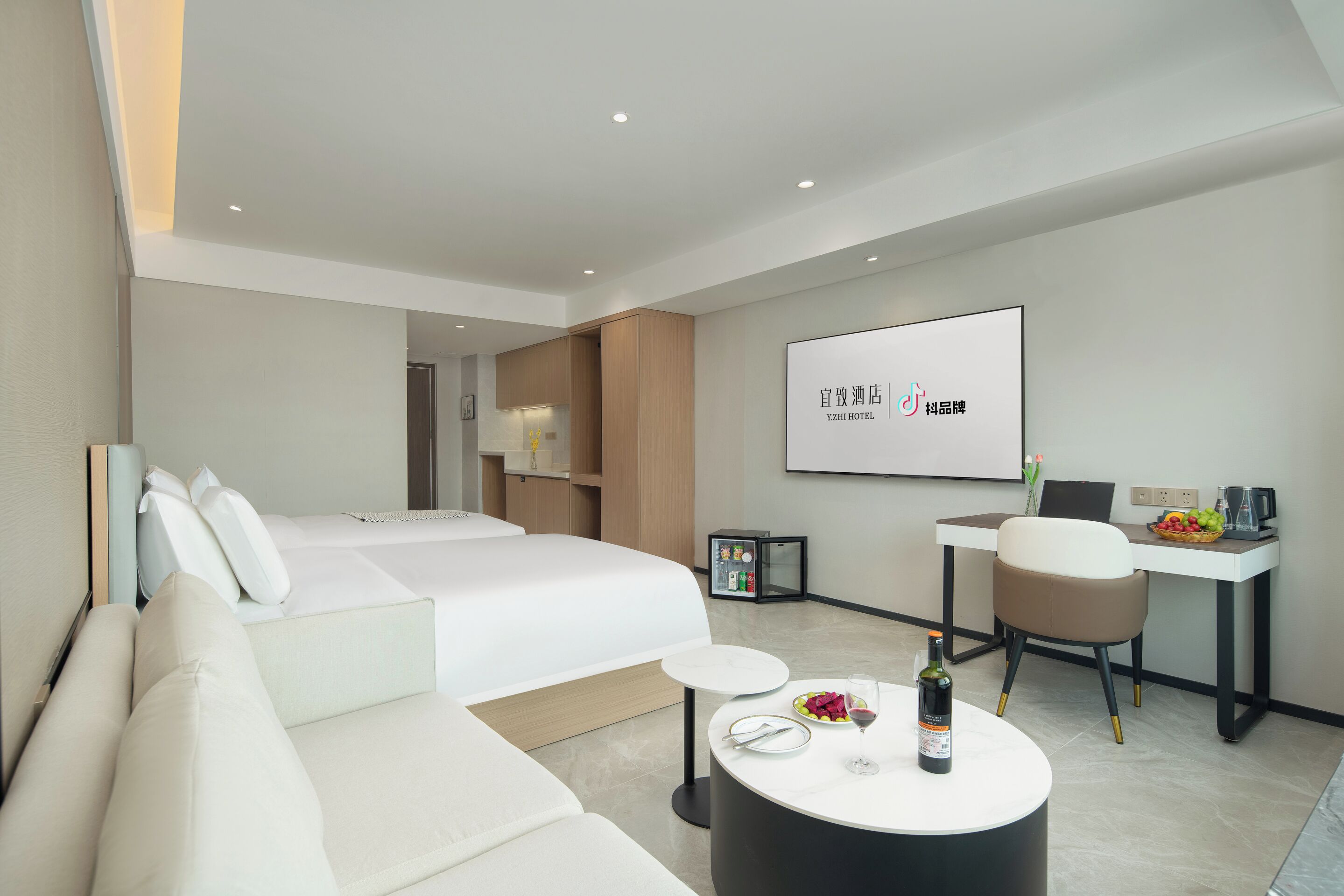 A modern hotel room with a bed, a sofa, a small round table with a bottle and glass, and a desk with a chair and a fruit bowl.