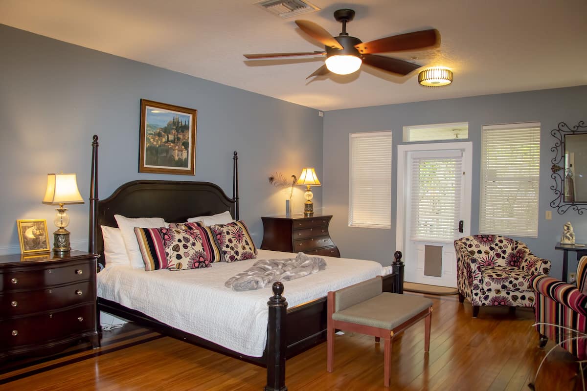 Une chambre avec un grand lit, deux tables de chevet avec des lampes, un ventilateur de plafond et un fauteuil.