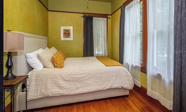 Un dormitorio con cama, mesita de noche, lámpara, un cuadro enmarcado en la pared y una ventana con cortinas.