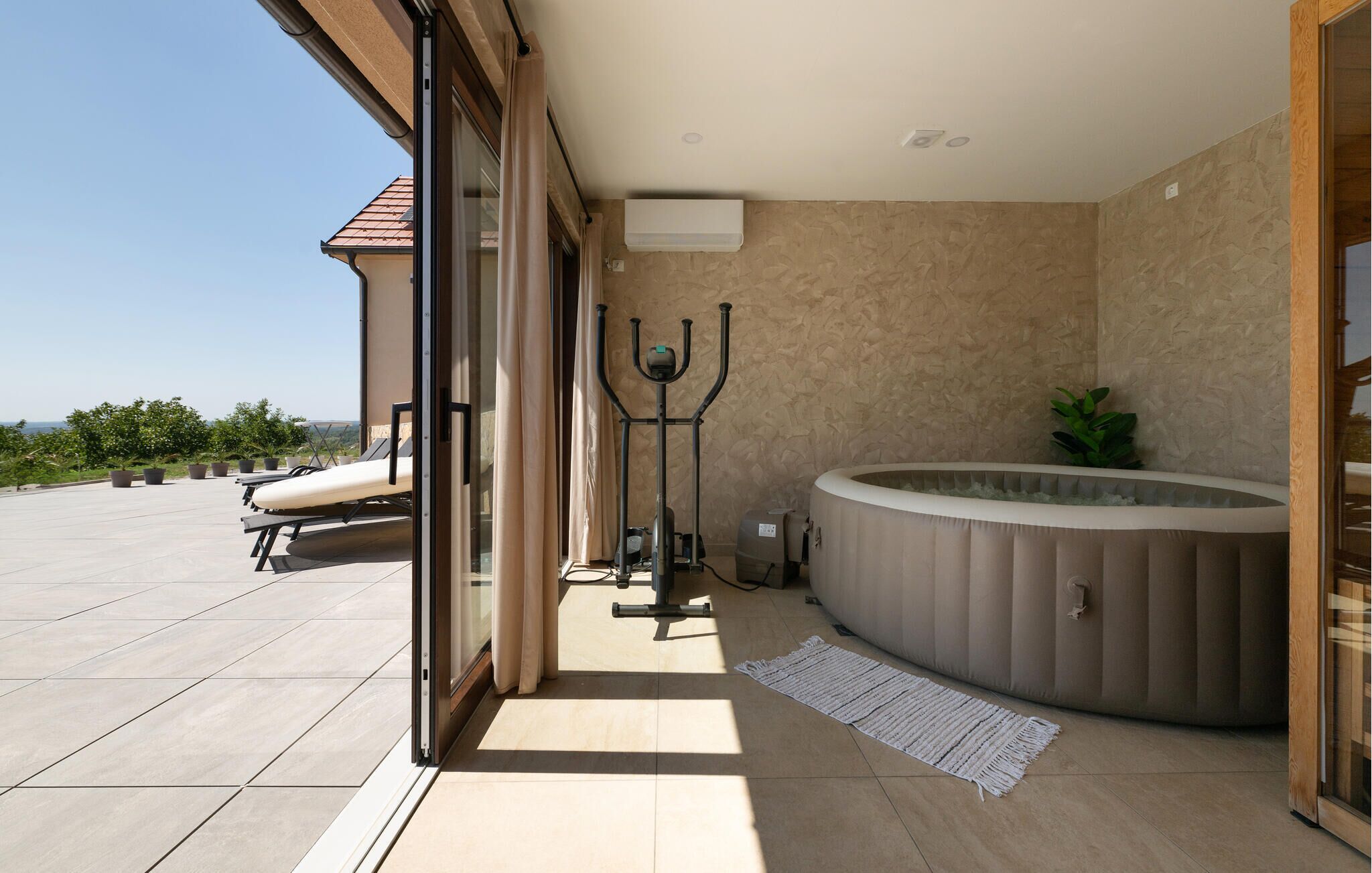 A modern bathroom with a bathtub, a large mirror, and a glass door leading to a balcony with sun loungers.