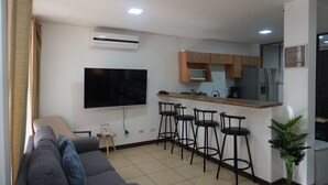 Un espacio habitable compacto con kitchenette, un televisor de pantalla plana montado en la pared, un sofá gris y una pequeña mesa redonda con un marco de fotos.