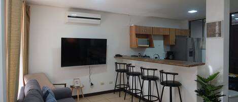 A compact living space with a kitchenette, a flat-screen TV mounted on the wall, a grey sofa, and a small round table with a picture frame.