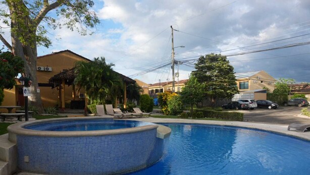 Una piscina con sillones de descanso, rodeada de palmeras y un edificio con techo de tejas.