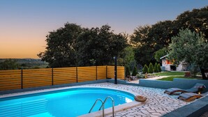 A swimming pool with a stone deck, a wooden fence, and a view of trees and a building in the background.