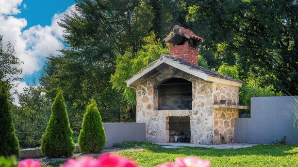 Una cocina al aire libre de piedra con chimenea, rodeada de césped y árboles.