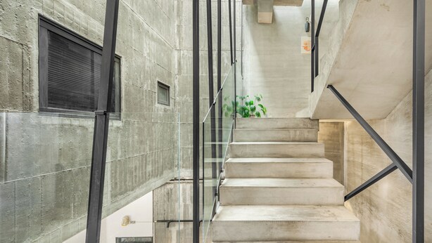 Un escalier moderne avec des rampes en verre et des murs en béton, menant à l’étage supérieur.