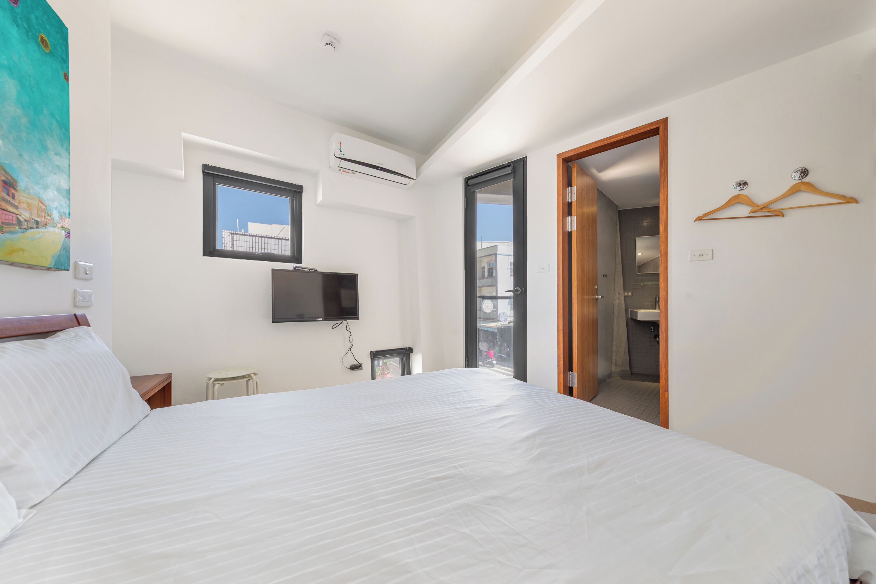 A hotel room with a large bed, a television, an air conditioning unit, a painting, and a view of the outside.