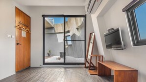 Une chambre moderne avec une porte en bois, une porte-fenêtre coulissante donnant sur un balcon, un banc en bois et un téléviseur fixé au mur.