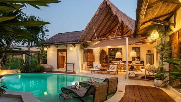 A poolside area with lounge chairs, a thatched-roof structure, and a well-lit indoor space with seating.