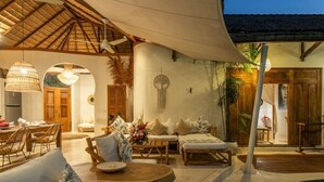 A cozy outdoor seating area with a round wooden table, cushioned chairs, and a sofa. There is a pool in the background, and the area is covered by a thatched roof.