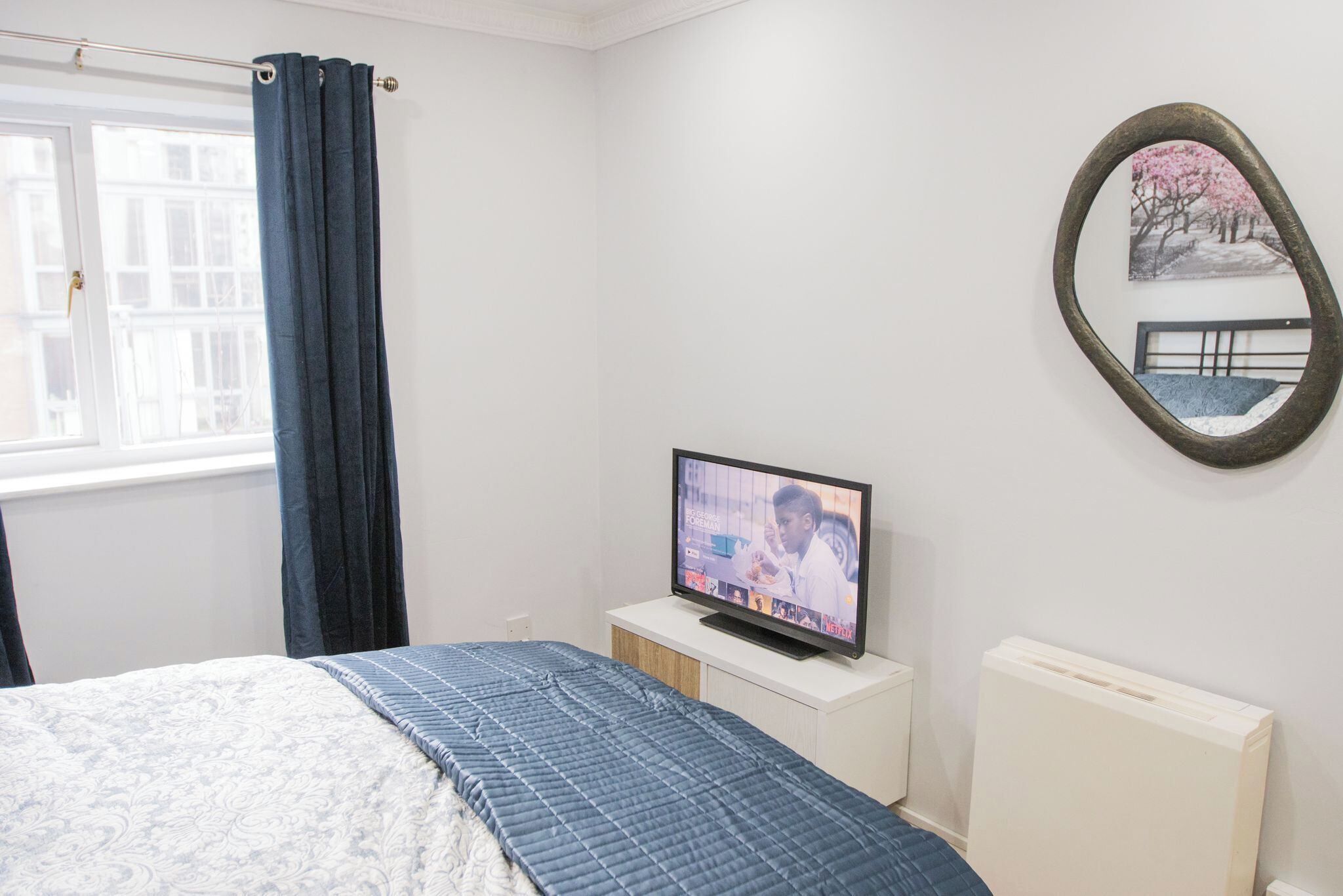 Un dormitorio con una cama, un televisor sobre una mesita de noche blanca, una ventana con cortinas y un espejo montado en la pared.
