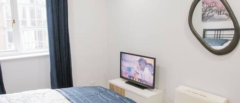 Un dormitorio con una cama, un televisor sobre una mesita de noche blanca, una ventana con cortinas y un espejo montado en la pared.